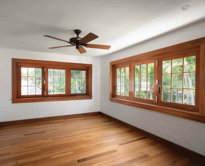 Empty room with hardwood floors, white walls, and wood-framed windows. A ceiling fan hangs from the ceiling.