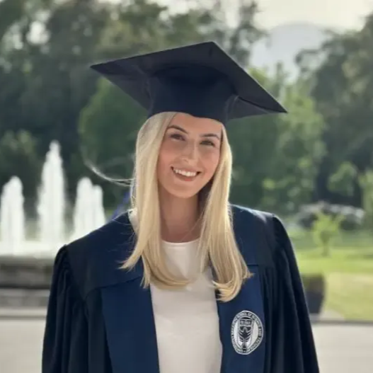 Kvinne i konfirmasjonslue og -kjole smiler foran en fontene.