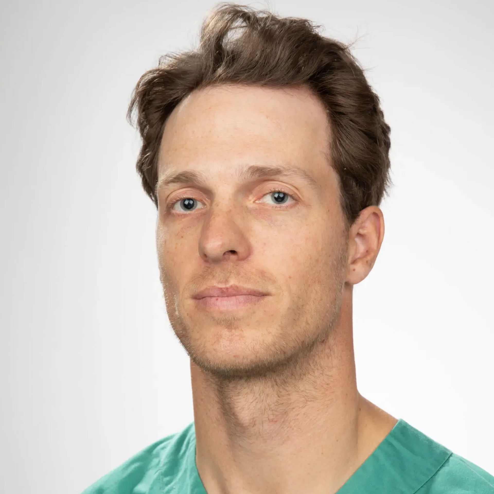 Mann i grønne skrubber ser direkte inn i kameraet med et nøytralt uttrykk mot en hvit bakgrunn.