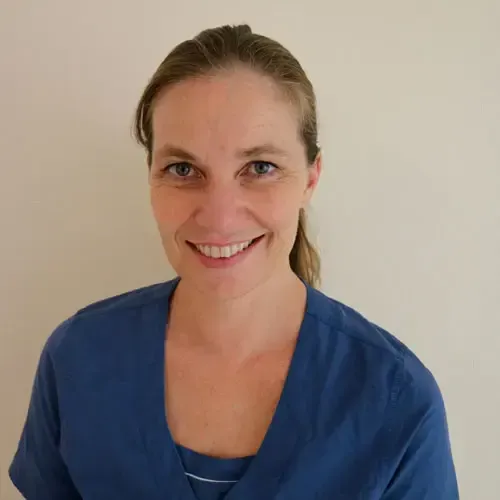 Kvinne i blå skrubber smiler foran en enkel vegg.