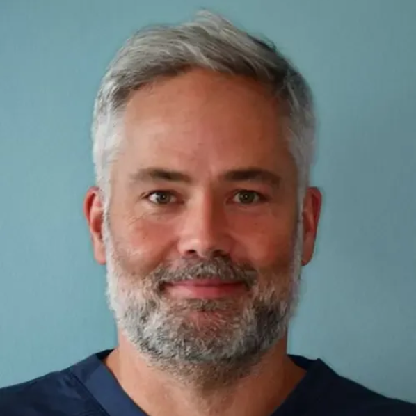 Mann med grått hår og skjegg smiler, iført en mørkeblå topp, mot en blå bakgrunn.