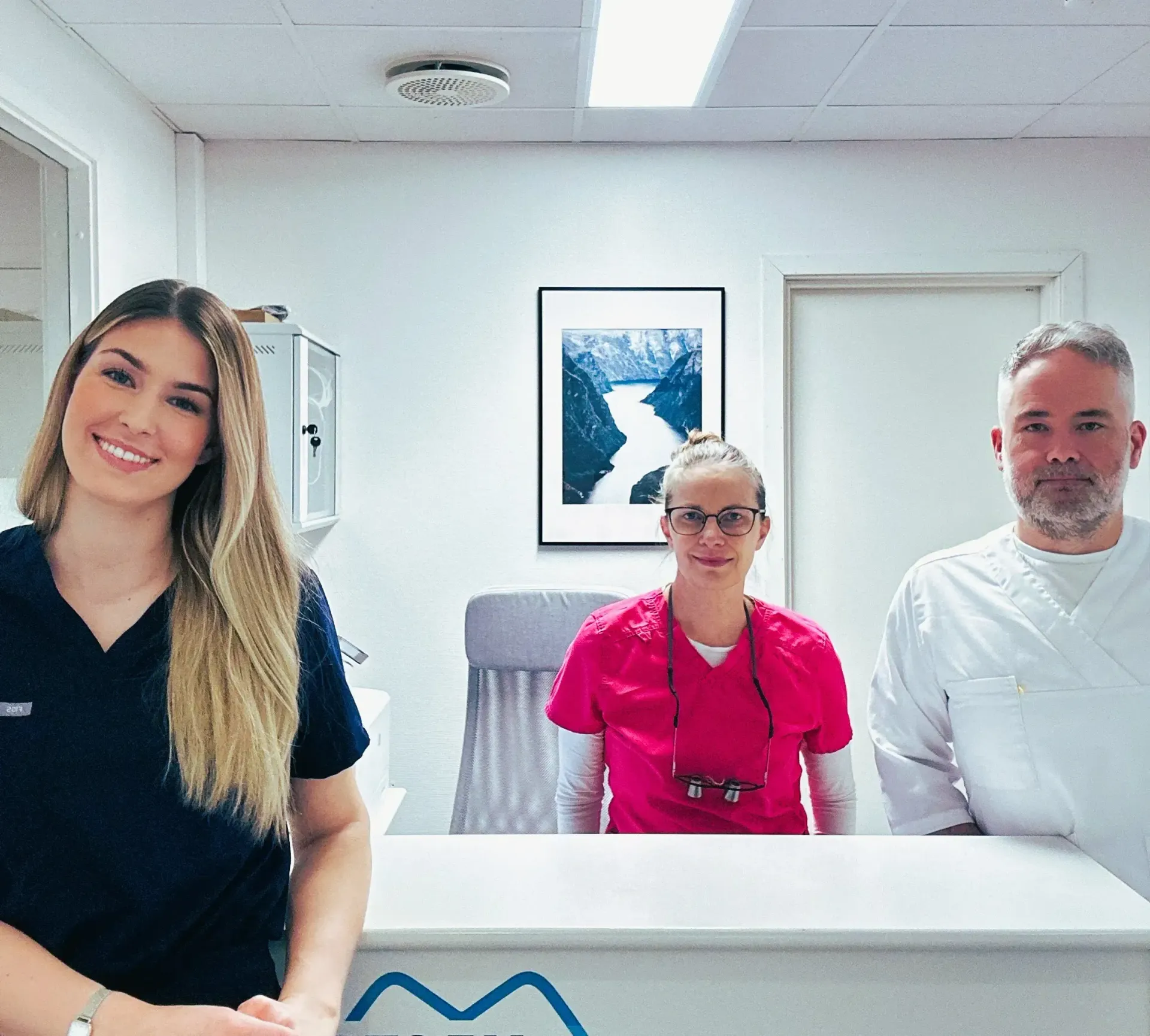 Tannlegekontorpersonalet bak en resepsjonsskranke: kvinne i marineblått, en annen i rosa og en mann i hvitt.