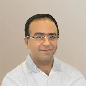 Mann med briller smiler, iført en lyseblå skjorte med krage. Ensfarget bakgrunn.