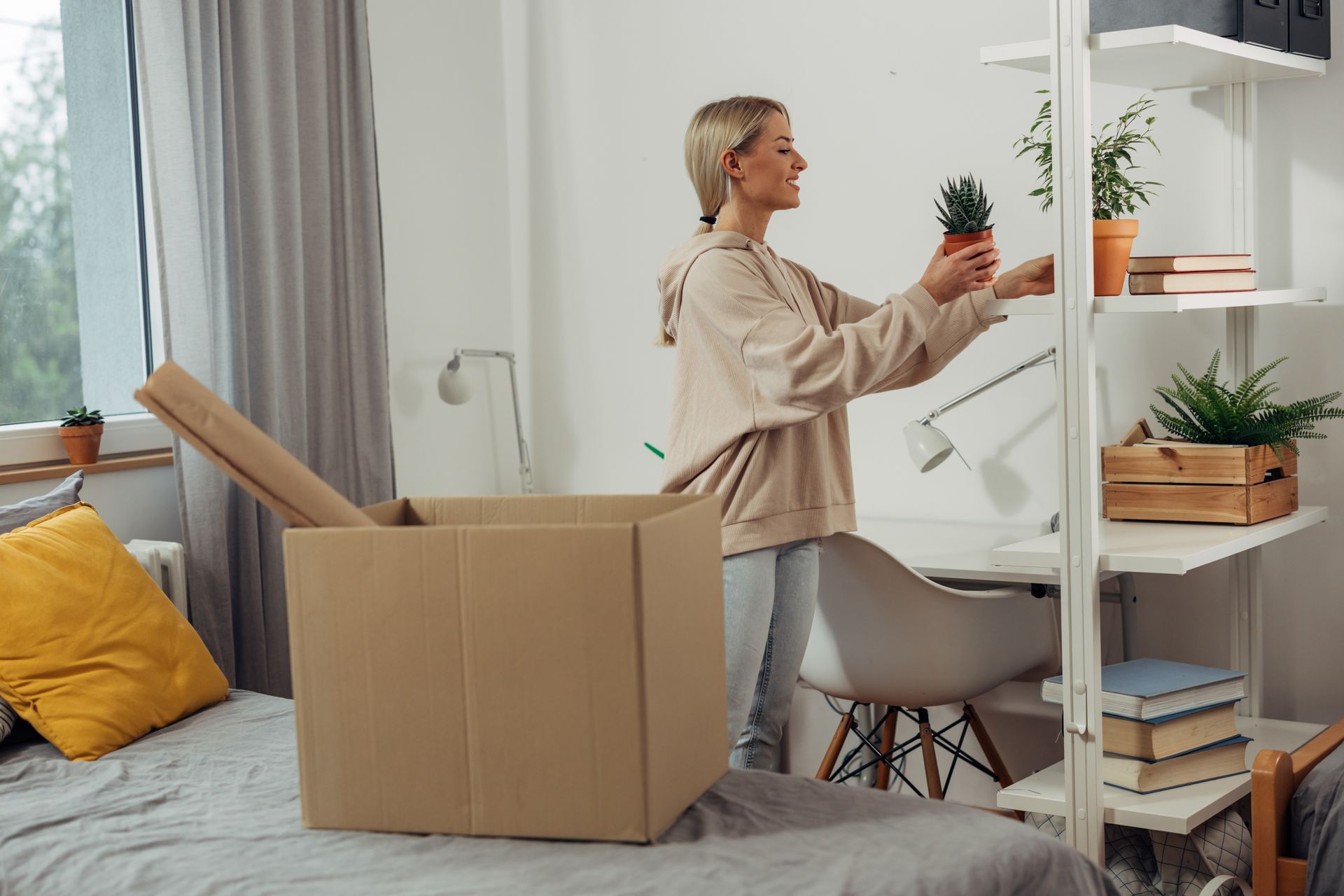 A person in a beige hoodie unpacks items, placing a small potted plant on a white shelving unit in a furnished bedroom.