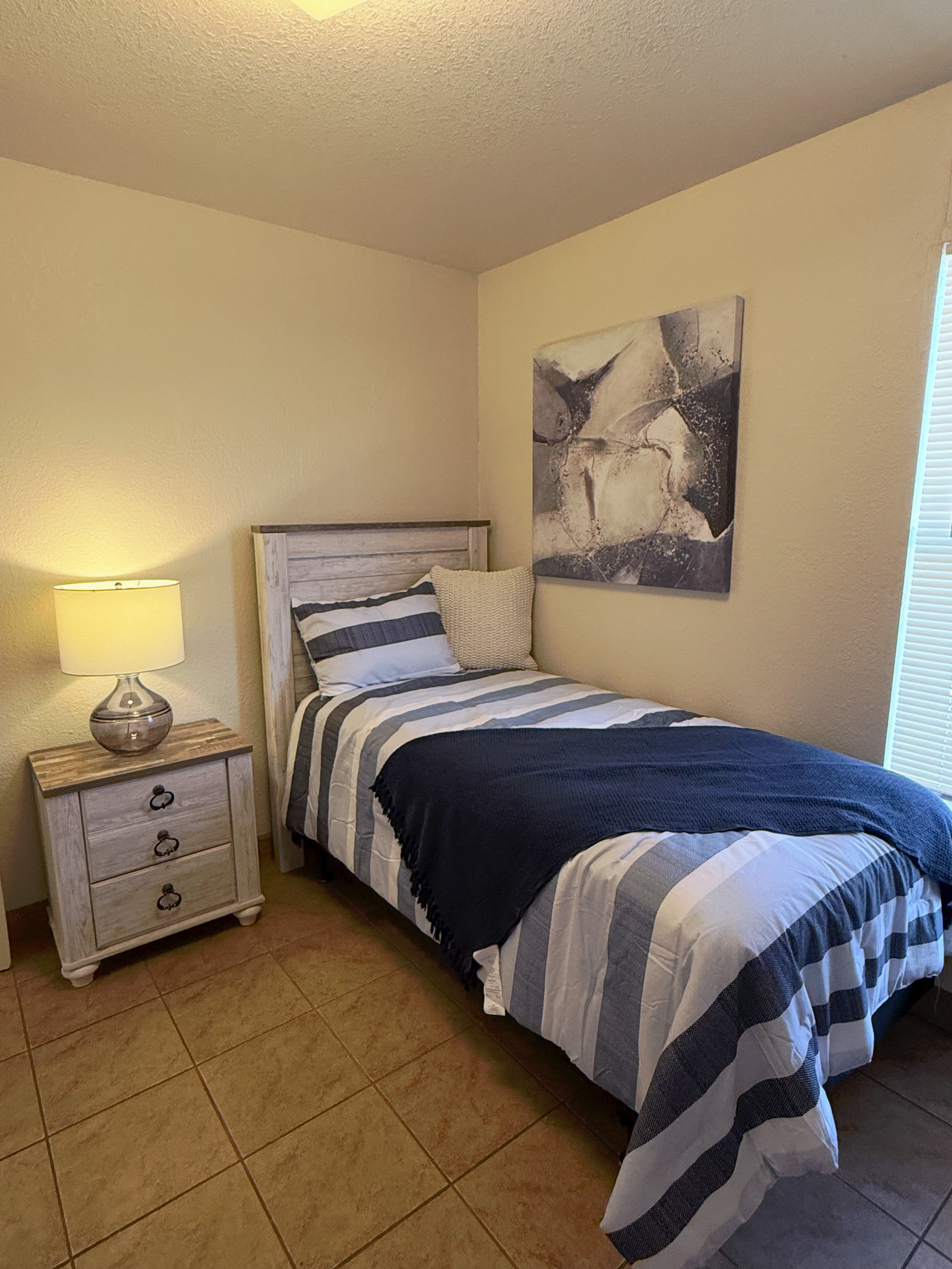 A small bedroom featuring a single bed with striped blue and white bedding, a white nightstand, and abstract wall art.