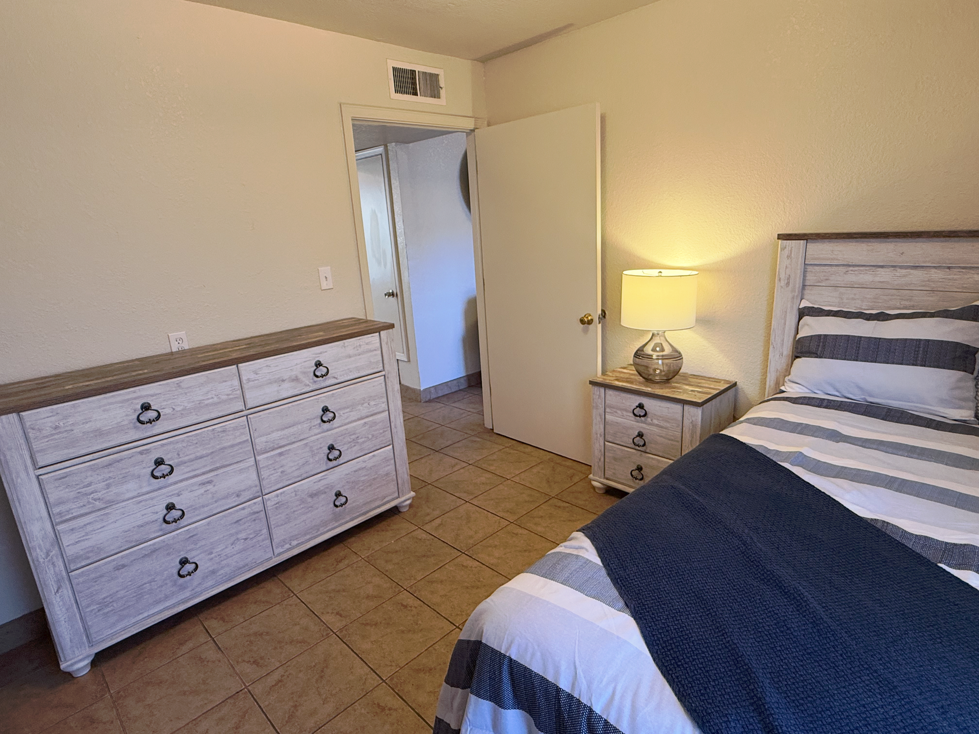 A bedroom with a bed featuring blue and white striped bedding, a matching white wood dresser, and a lit bedside lamp.
