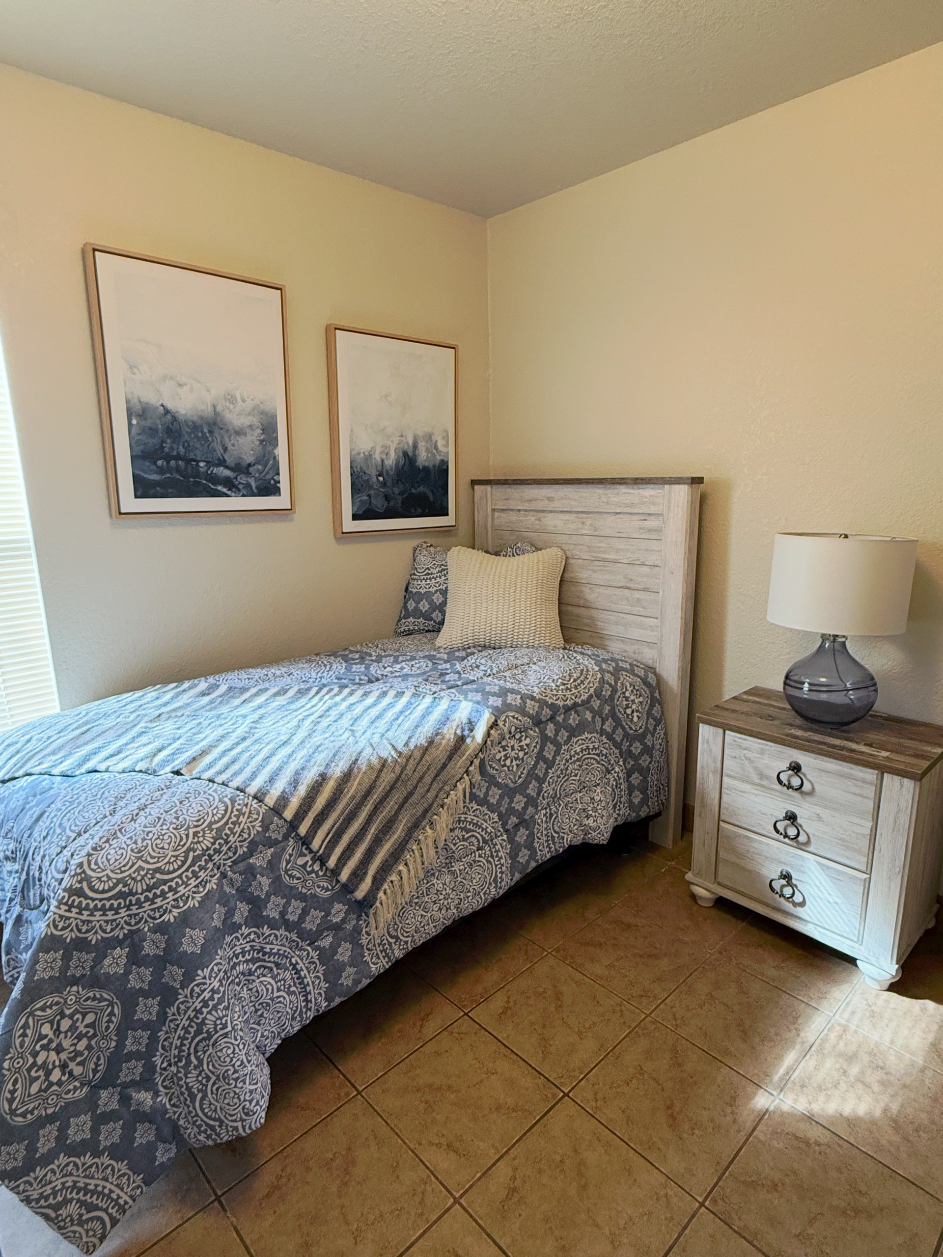 A twin bed with blue-and-white patterned bedding, next to a white nightstand with a lamp and two abstract blue wall prints.