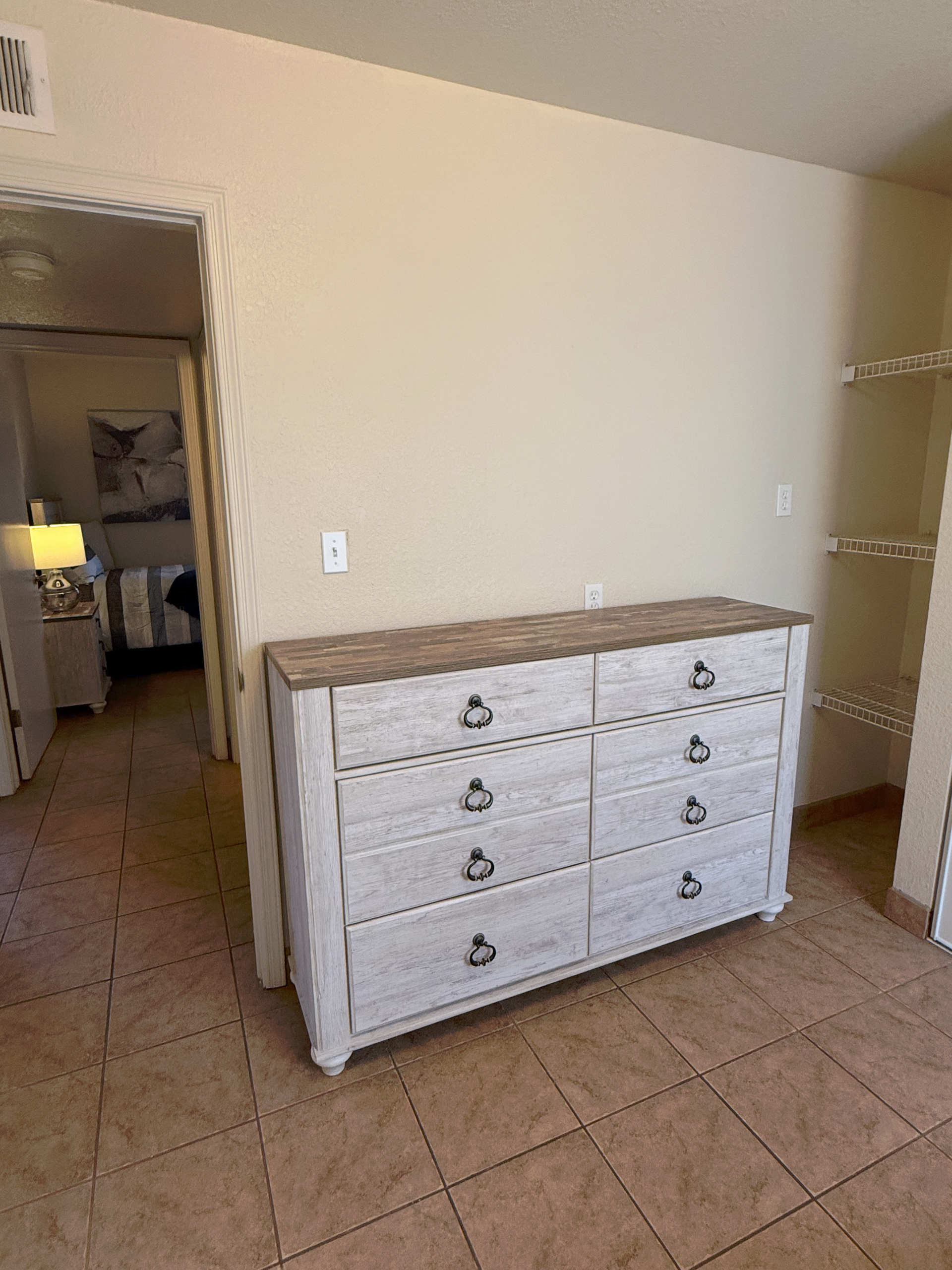 A white, weathered-wood dresser with eight drawers stands against a beige wall in a room with tiled floors.