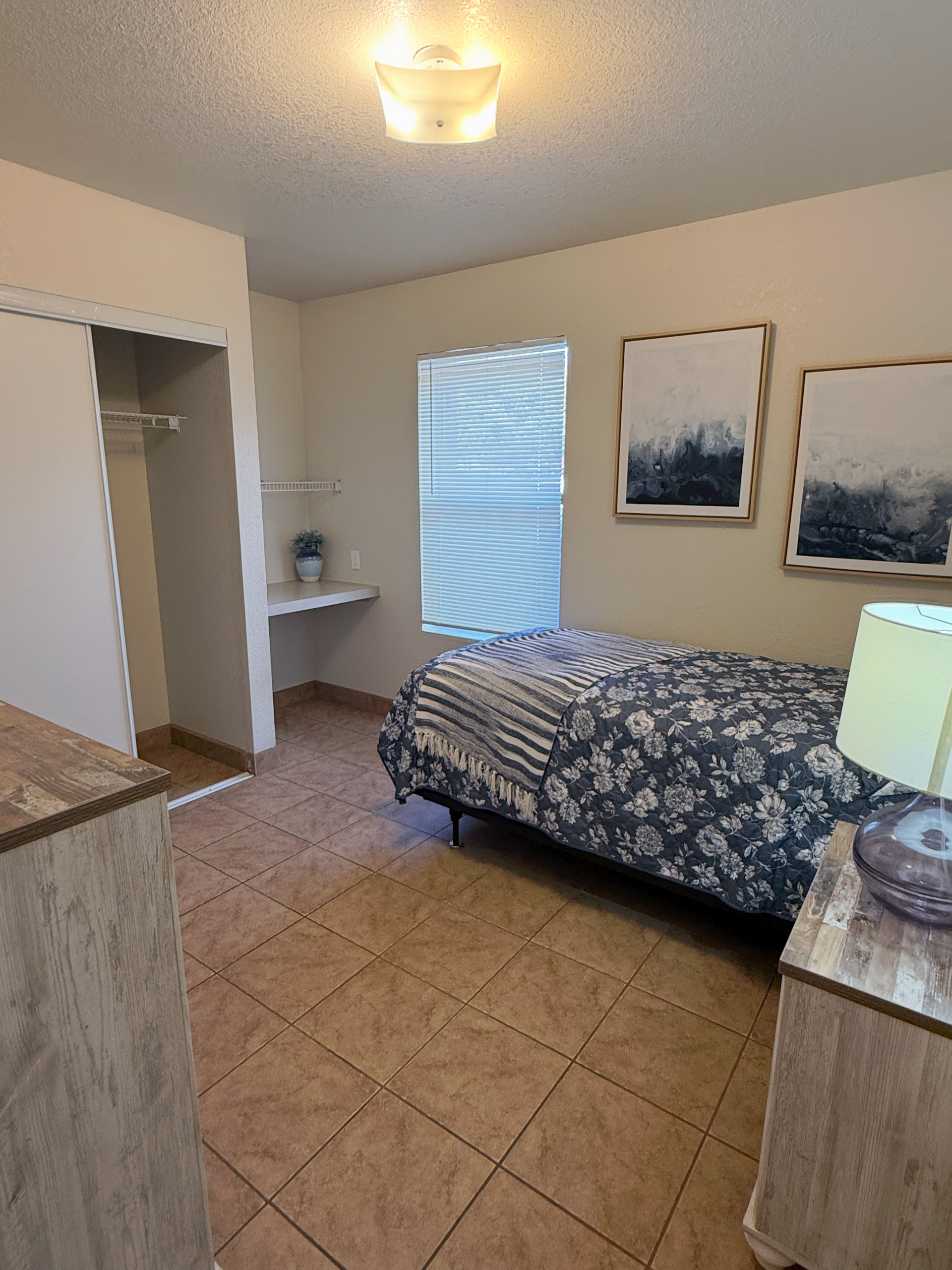 A bedroom with tan tiled floors, a single bed with a floral quilt, two wall paintings, and a built-in desk by the window.