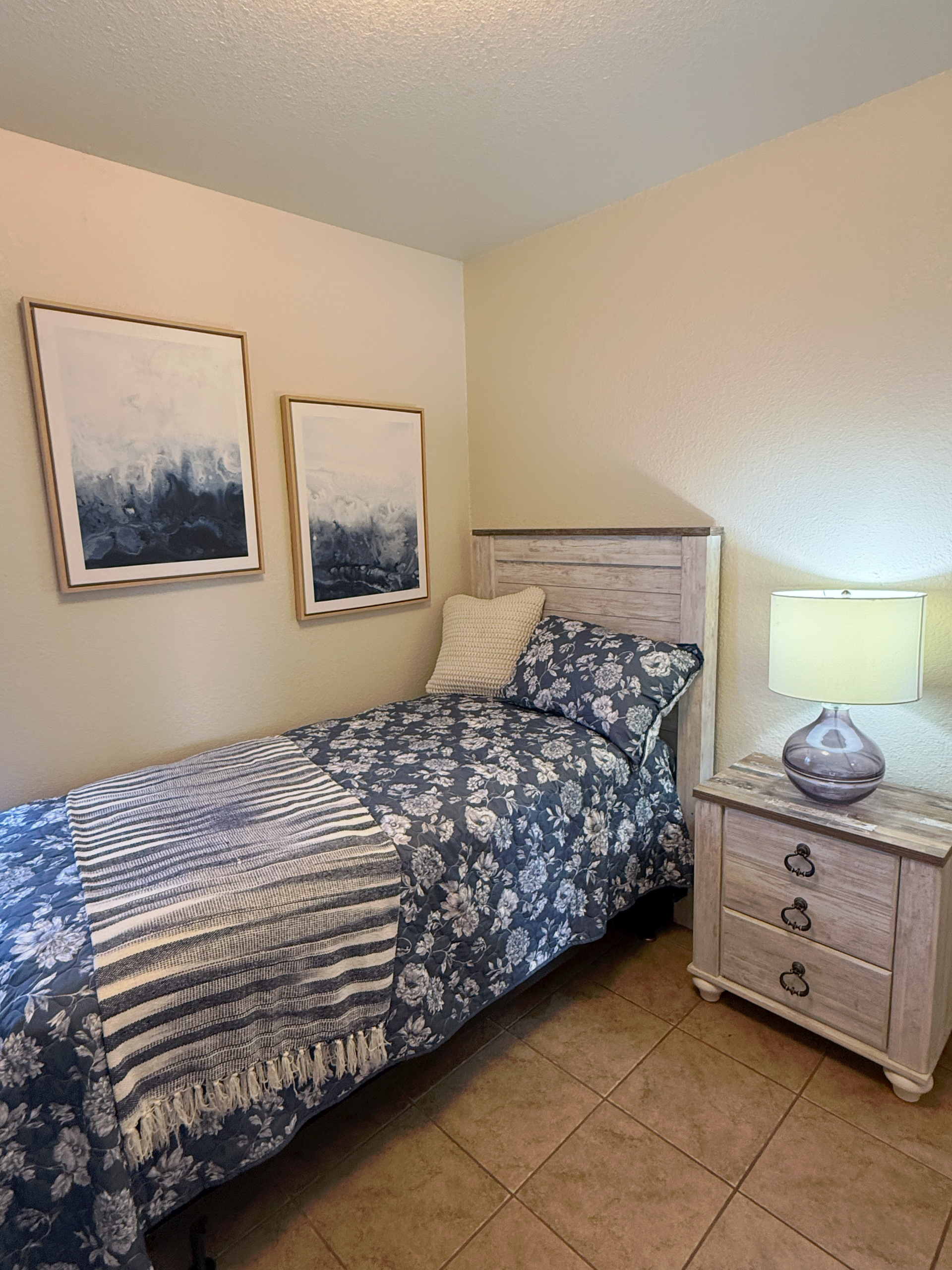 A bedroom with a single bed featuring blue and white floral bedding, a matching throw, and two framed abstract artworks.