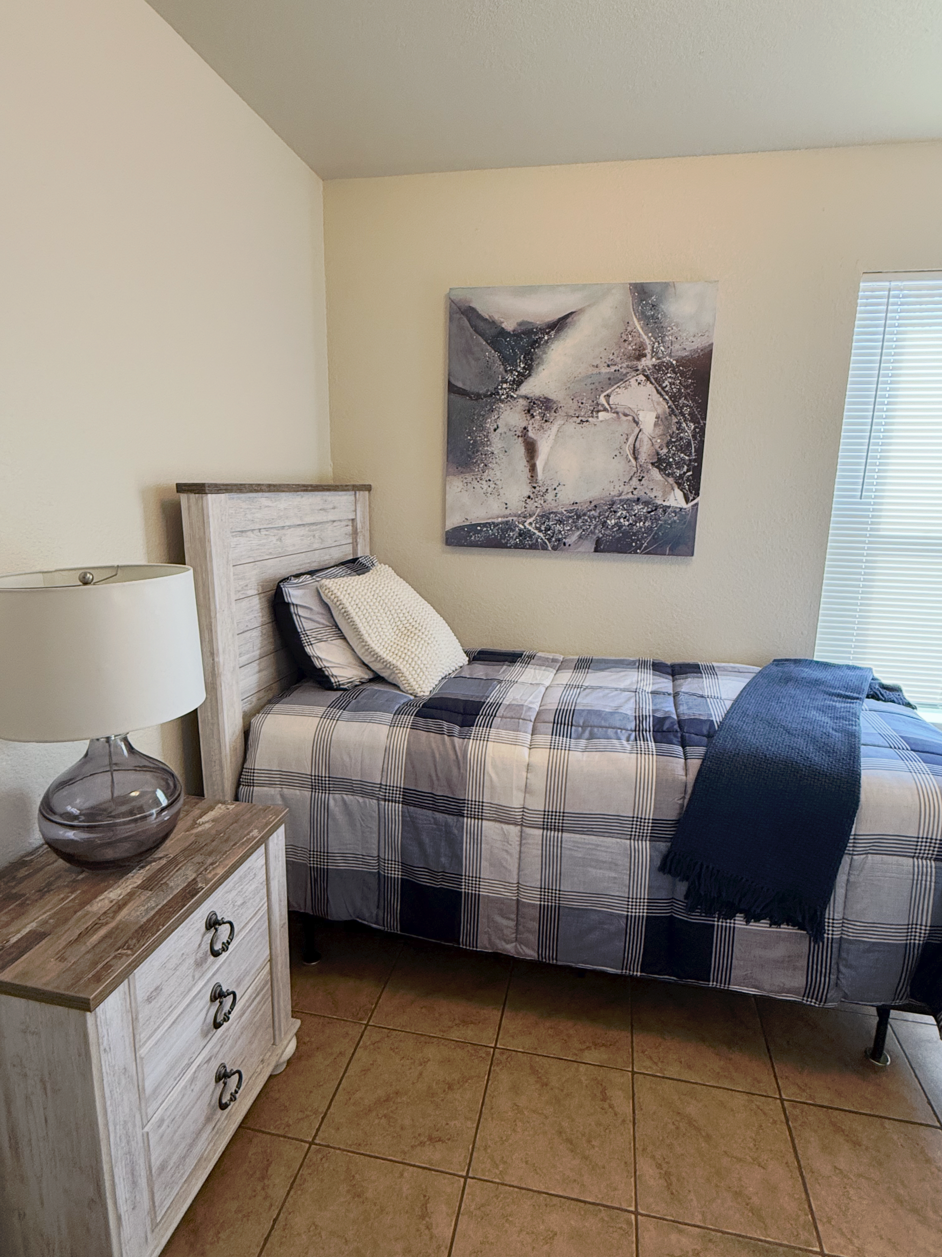 A bedroom with a rustic white nightstand, lamp, a single bed with blue plaid bedding, and abstract wall art.