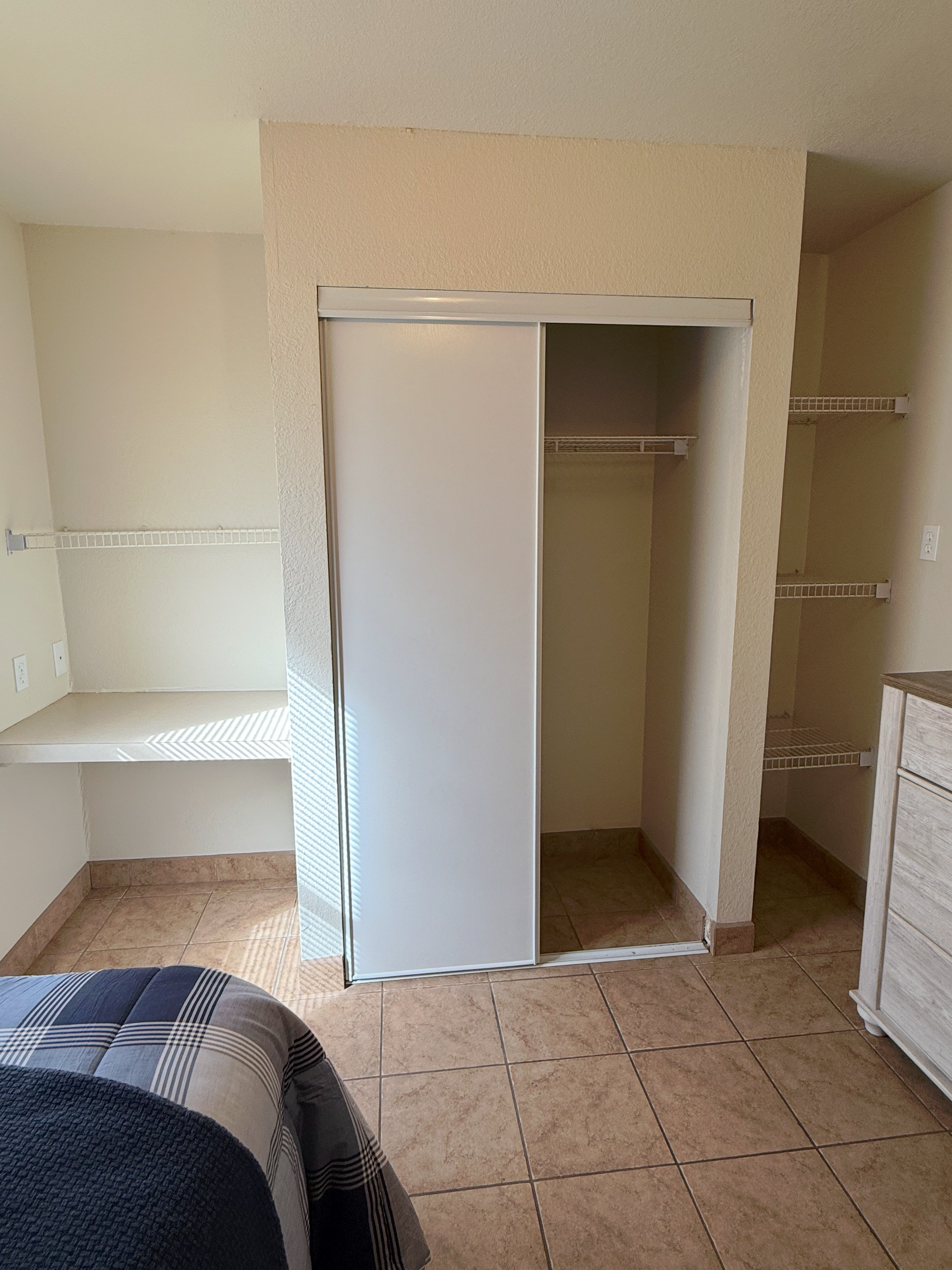 A bedroom interior featuring a closet with sliding white doors, open shelving to the left and right, and tiled flooring.