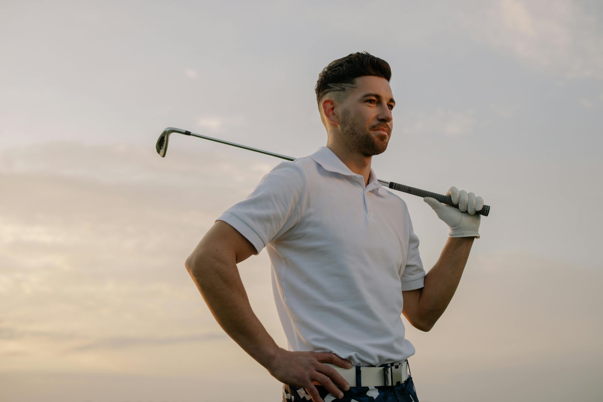 Man in white polo shirt and golf glove, holding a golf club on shoulder, looking away.