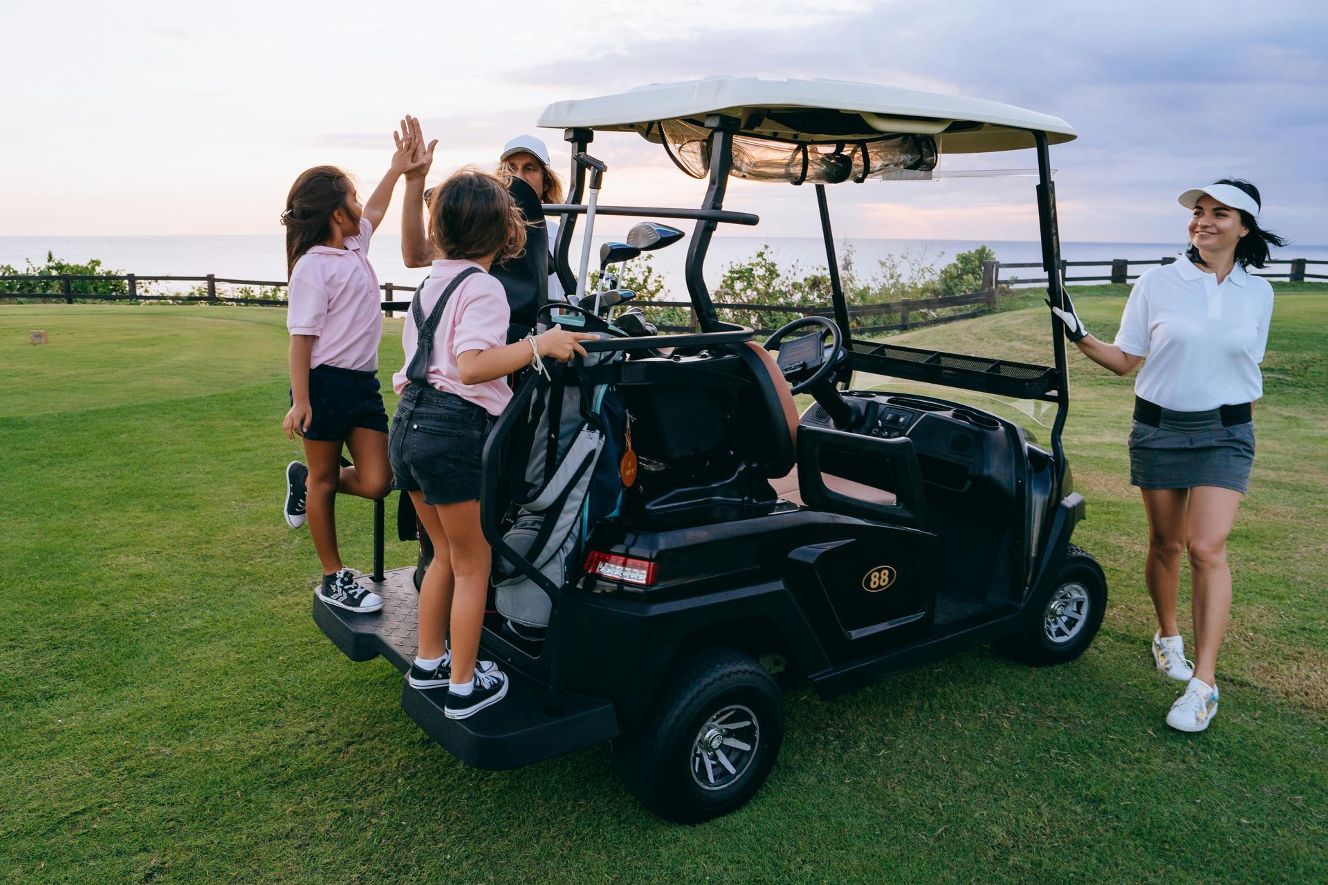 Family high-fives near a golf cart on a green course by the ocean.