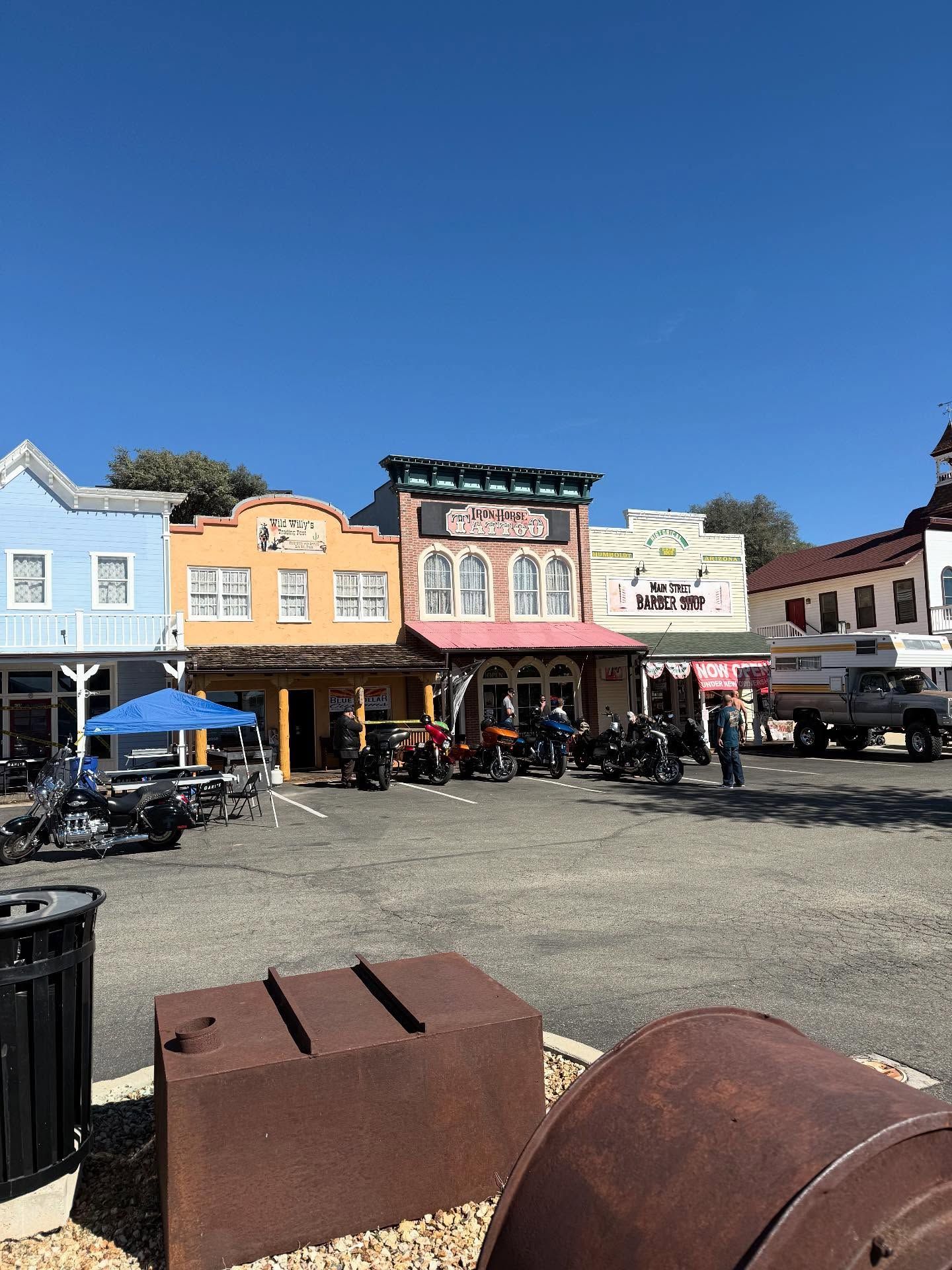 A row of colorful rustic-style storefronts under a blue sky, with several motorcycles parked in the gravel lot out front.