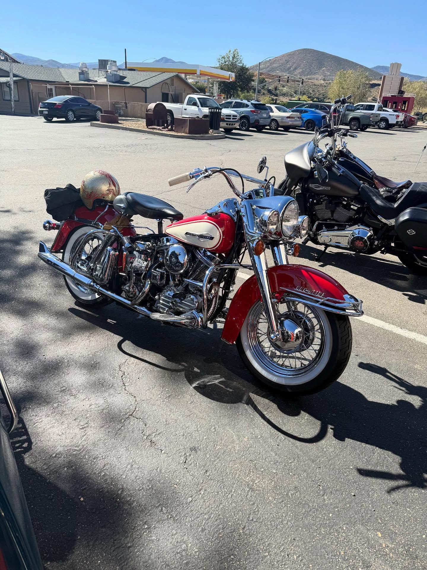 A red and cream vintage-style motorcycle parked on an asphalt lot with mountains and buildings in the background.