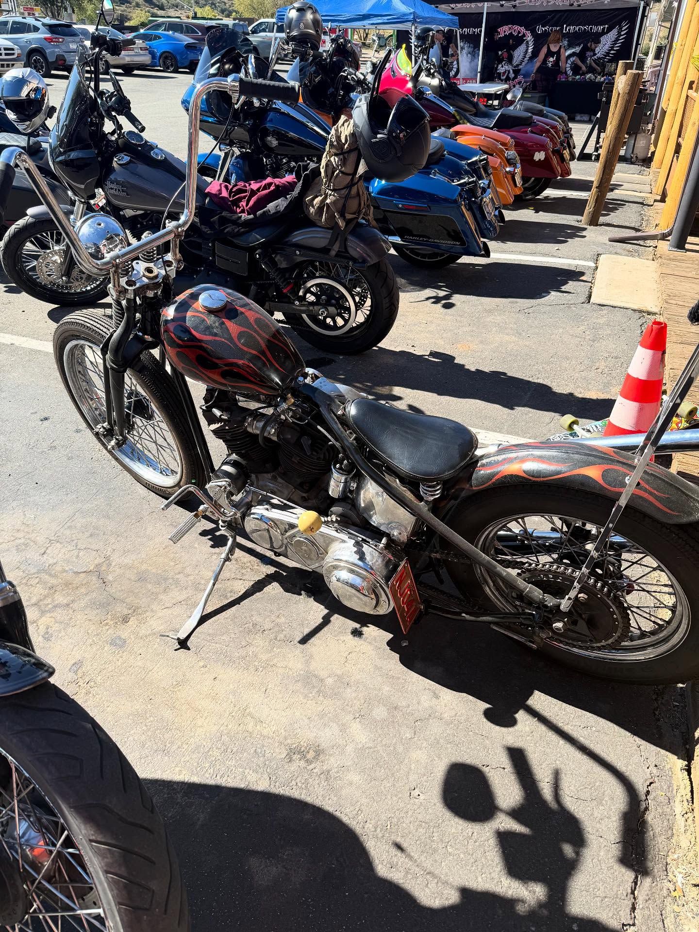 A custom, dark-painted bobber motorcycle parked on pavement in an outdoor lot filled with other motorcycles.