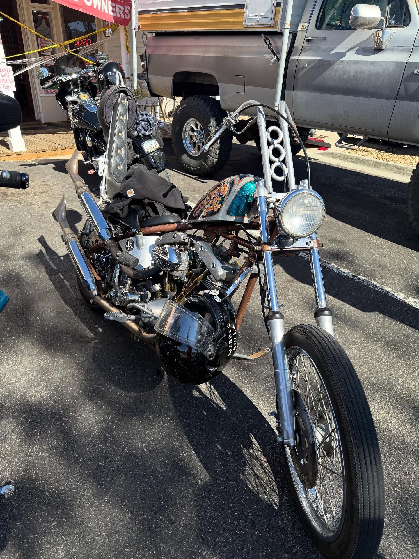 A turquoise custom chopper motorcycle parked outdoors in a sunlit lot, angled toward the camera with chrome details.