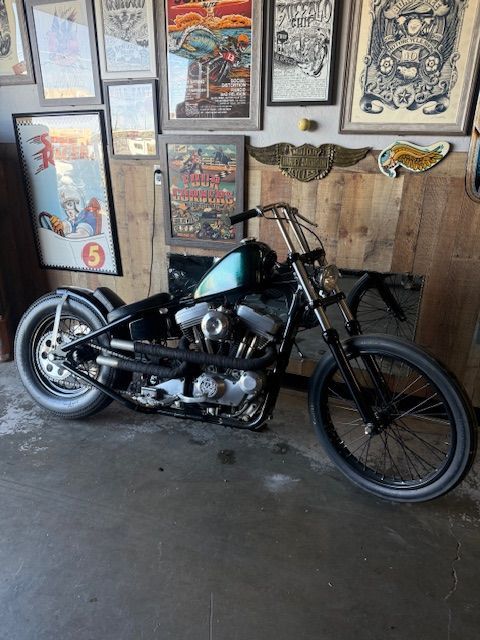 A black custom chopper motorcycle with a dark green tank parked inside a room decorated with wall-mounted posters.
