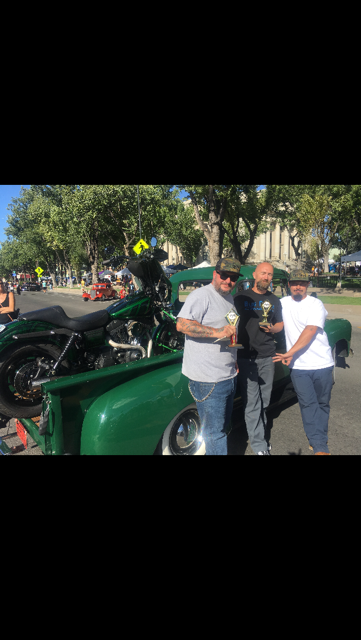 Three people stand with trophies in front of a green pickup truck carrying a motorcycle at an outdoor event.