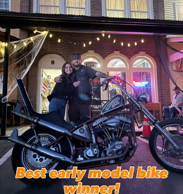 A couple poses with a custom chopper motorcycle in front of a brick building decorated with string lights.