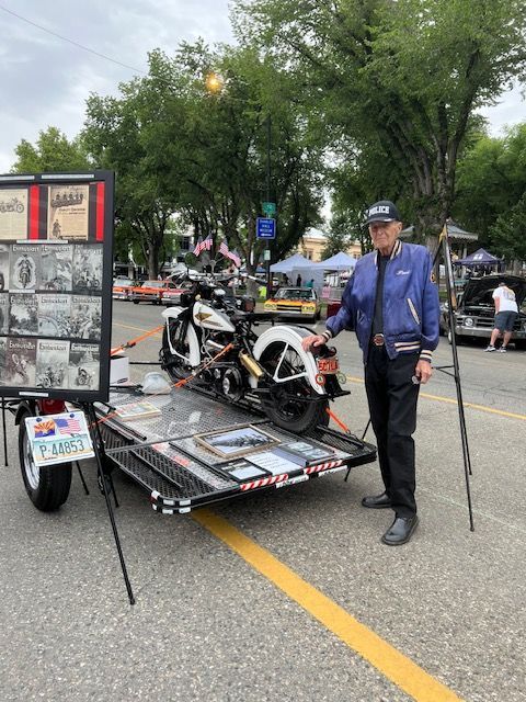 A person in a blue jacket stands beside a vintage motorcycle displayed on a trailer at an outdoor car show.
