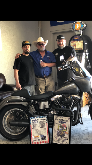 Three people stand with a custom motorcycle, holding two framed event posters in front of a vintage slot machine.