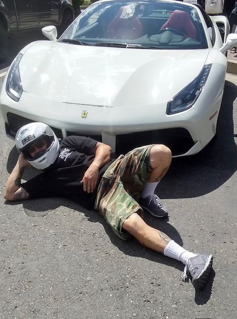 A person wearing a white helmet reclines on the asphalt in front of a white Ferrari.