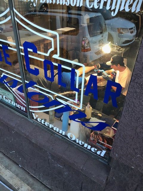 Through a glass window, a tattoo artist works on a client in a Blue Collar Tattoos shop, which is veteran-owned.
