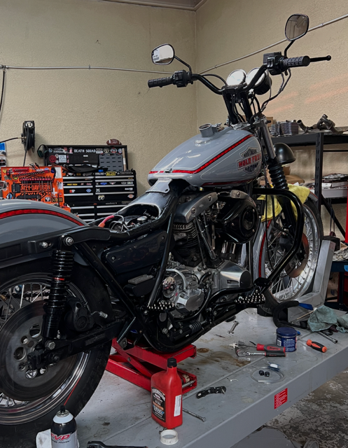A light gray Harley-Davidson motorcycle under repair, elevated on a red stand inside a cluttered garage workshop.