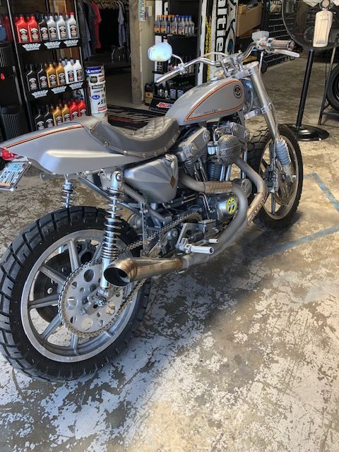 A custom silver Harley-Davidson Sportster motorcycle parked inside a workshop with rows of oil bottles in the background.