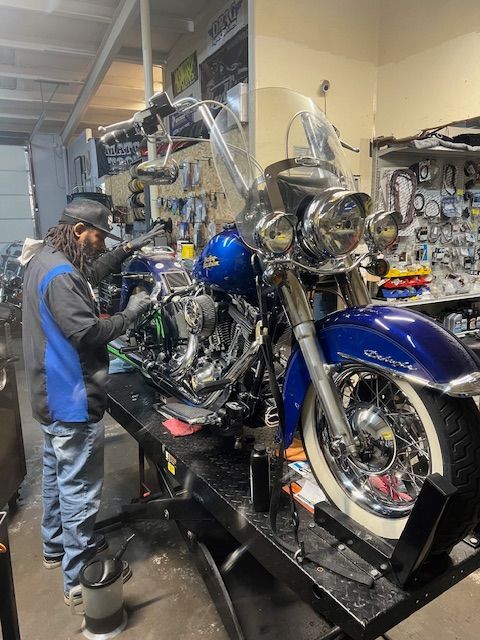 A mechanic in a blue and gray uniform works on a blue Harley-Davidson motorcycle on a lift in a repair shop.