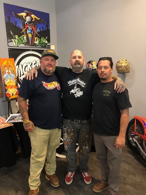 Three men pose together in a studio, with art, helmets, and a motorcycle visible in the background.