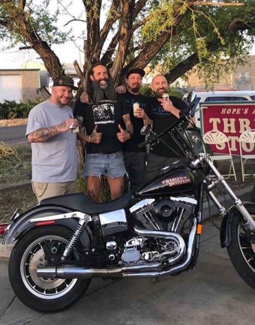 Four people stand smiling behind a parked black Harley-Davidson motorcycle in an outdoor, sunset setting.