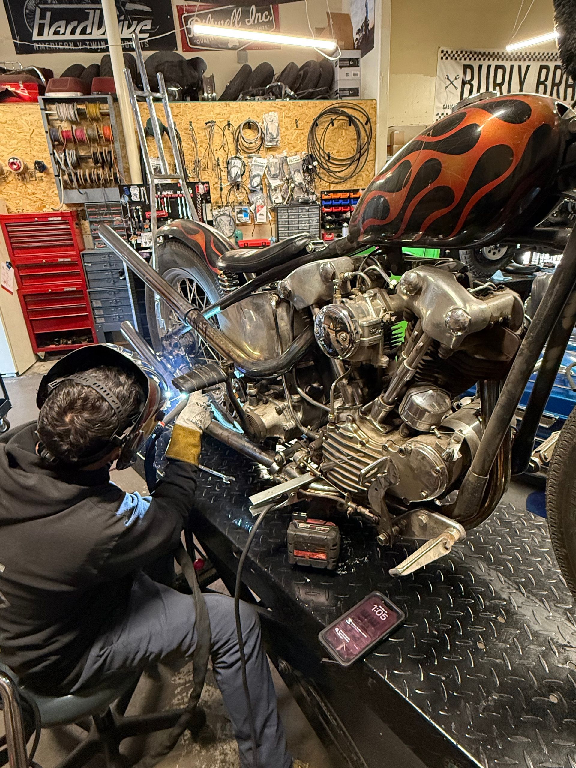 A person wearing a welding helmet works on a vintage-style motorcycle in a cluttered garage workshop.