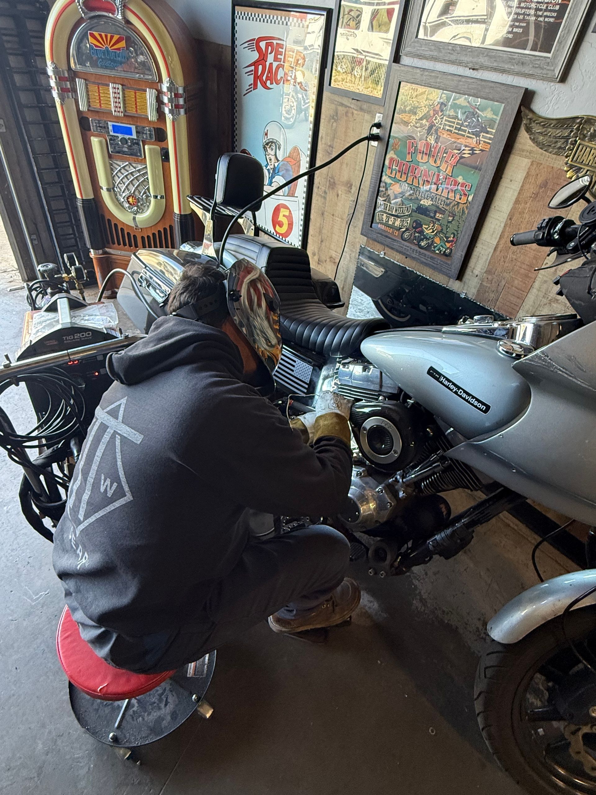 A person in a gray hoodie sits on a red stool, working on the engine of a silver motorcycle in a shop.
