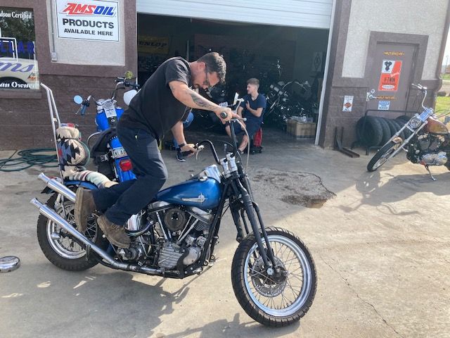 A person sits on a blue custom motorcycle in front of an open garage door, with another bike parked nearby.