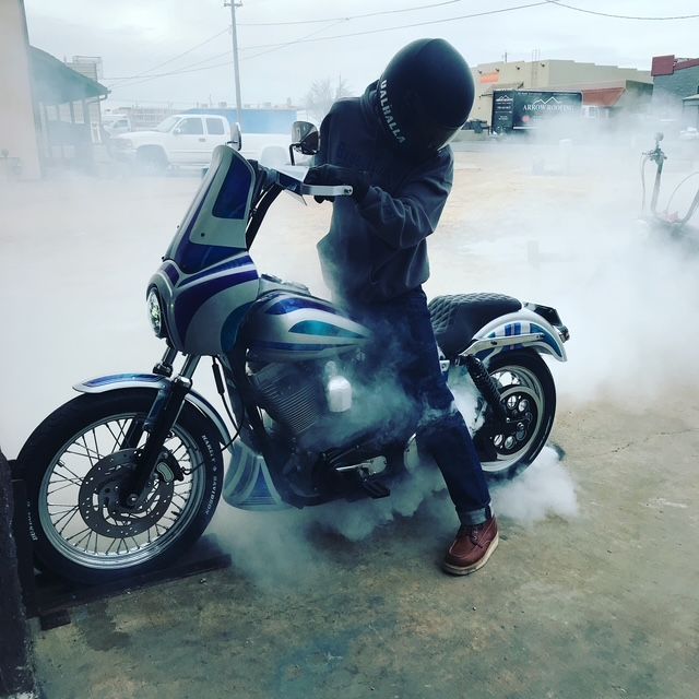 A person in a helmet sits on a blue and silver motorcycle, causing the rear tire to spin and produce a large cloud of smoke.