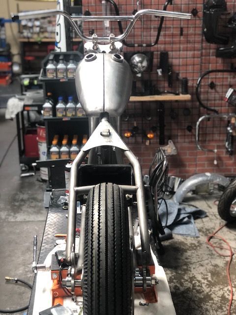 A bare-metal motorcycle frame with a fuel tank and handlebars, sitting on a lift in a workshop.