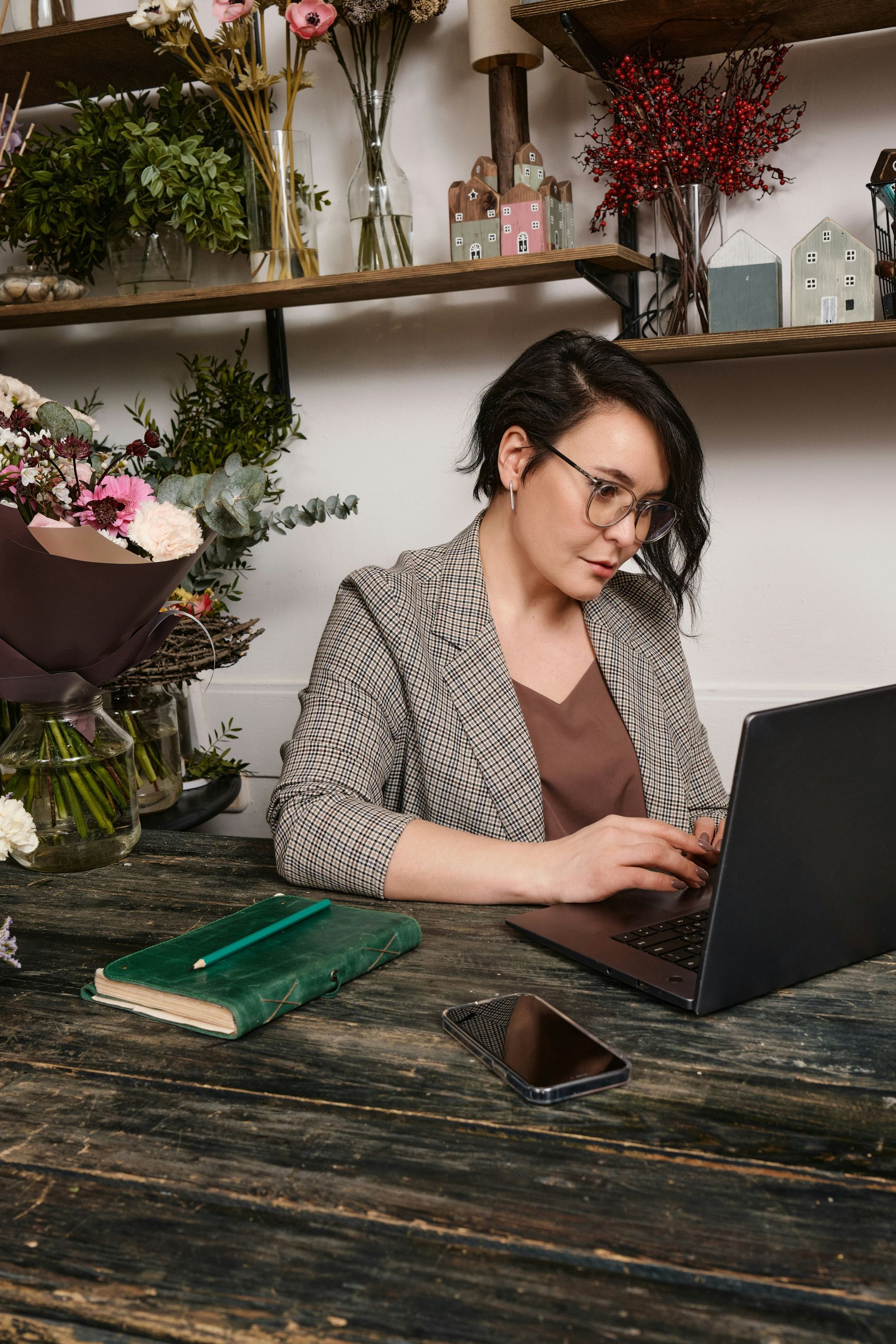 Woman in glasses and blazer types on laptop at a wooden table, notebook and phone nearby, with flowers in the background.