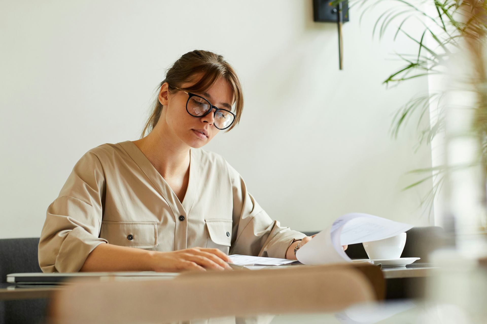 Woman with glasses reads papers at a table, next to a coffee cup and plant. Neutral background.