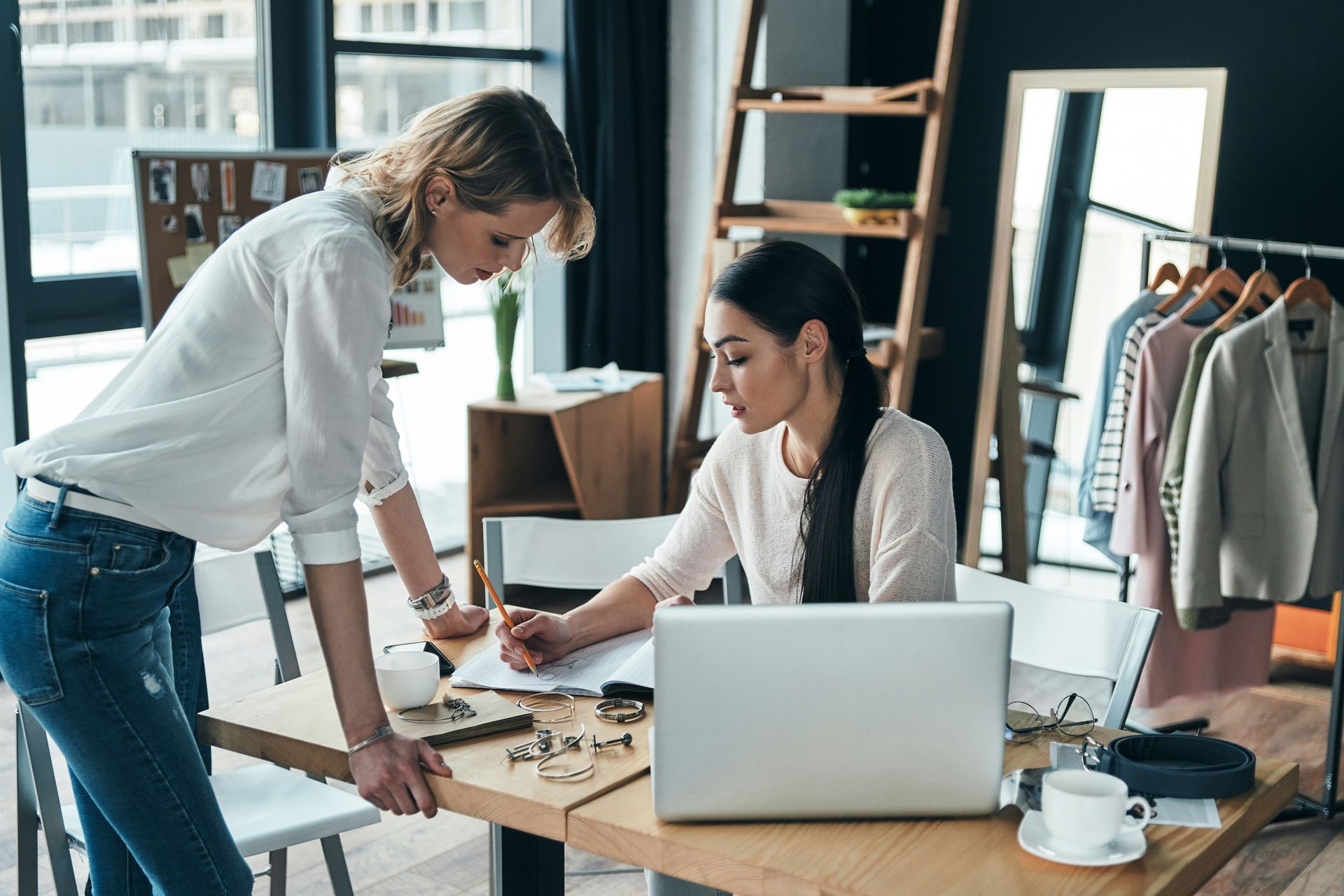 Two women collaborate at a desk with a laptop and papers in a bright office.