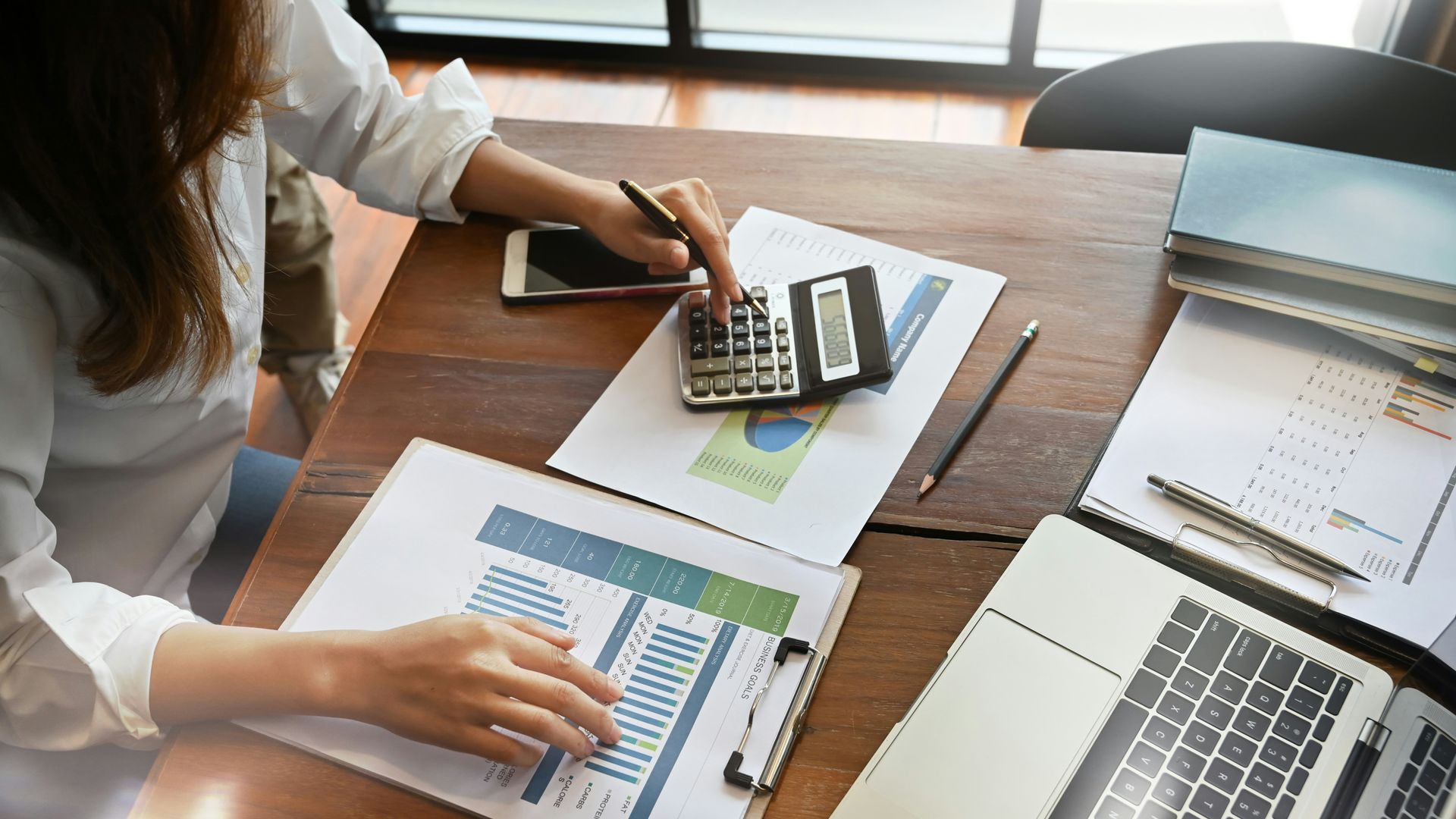 Woman calculating on a desk with papers, graphs, a calculator, phone, and laptop.