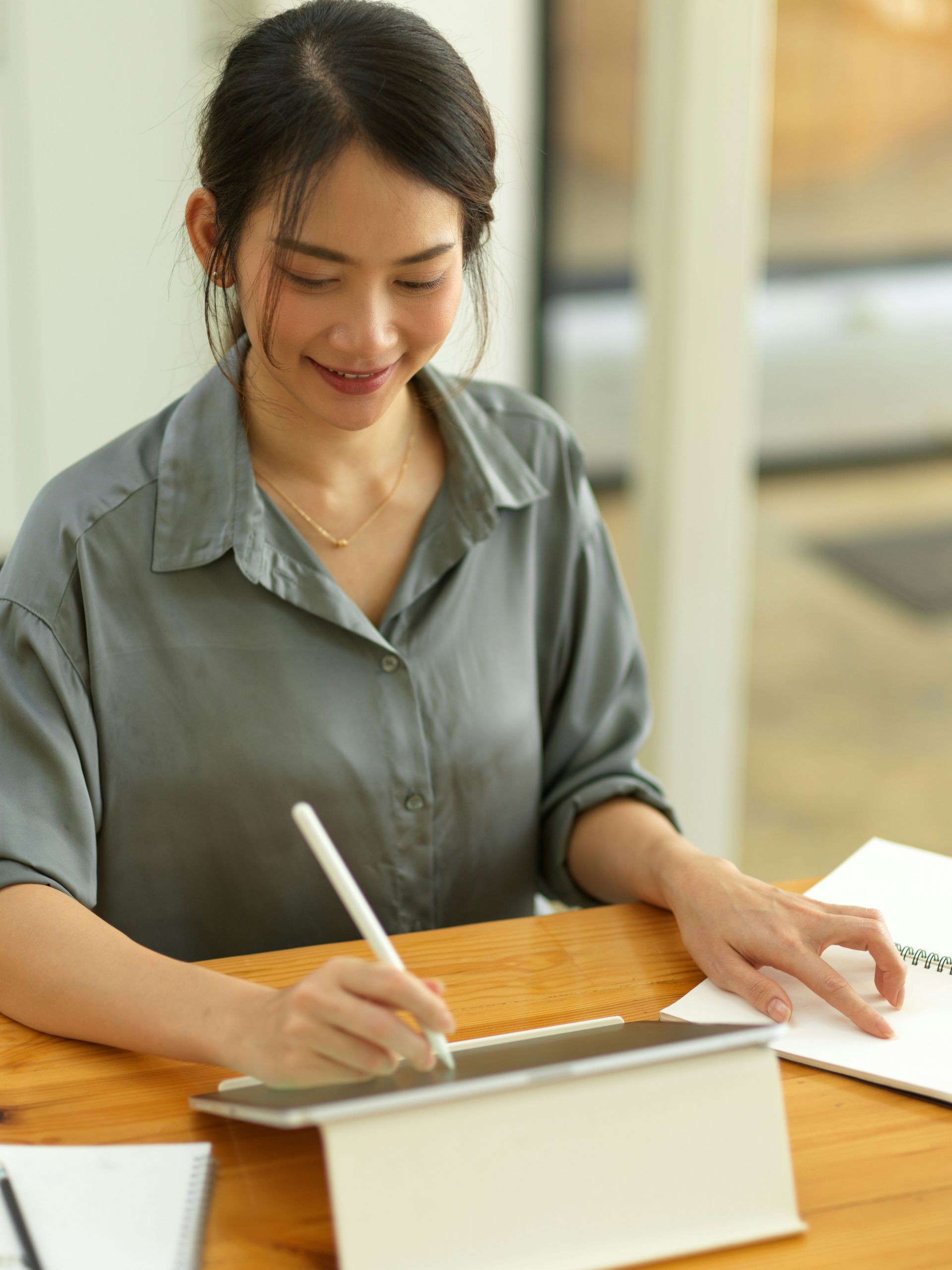 Woman smiles, writing on tablet with stylus at a wooden desk, notebook beside her.