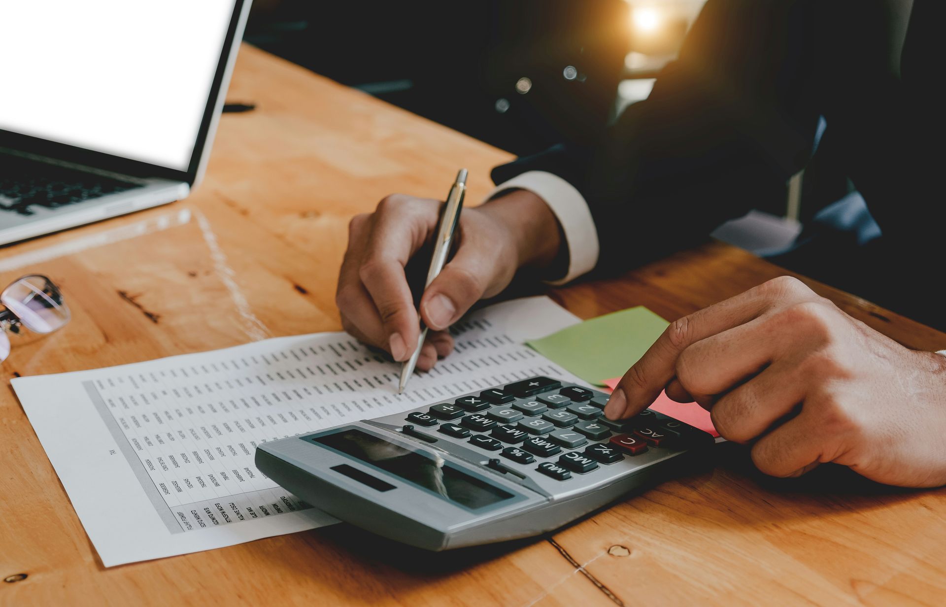 Person in a suit calculating on a calculator while looking at paperwork, with a laptop in the background on a wooden desk.