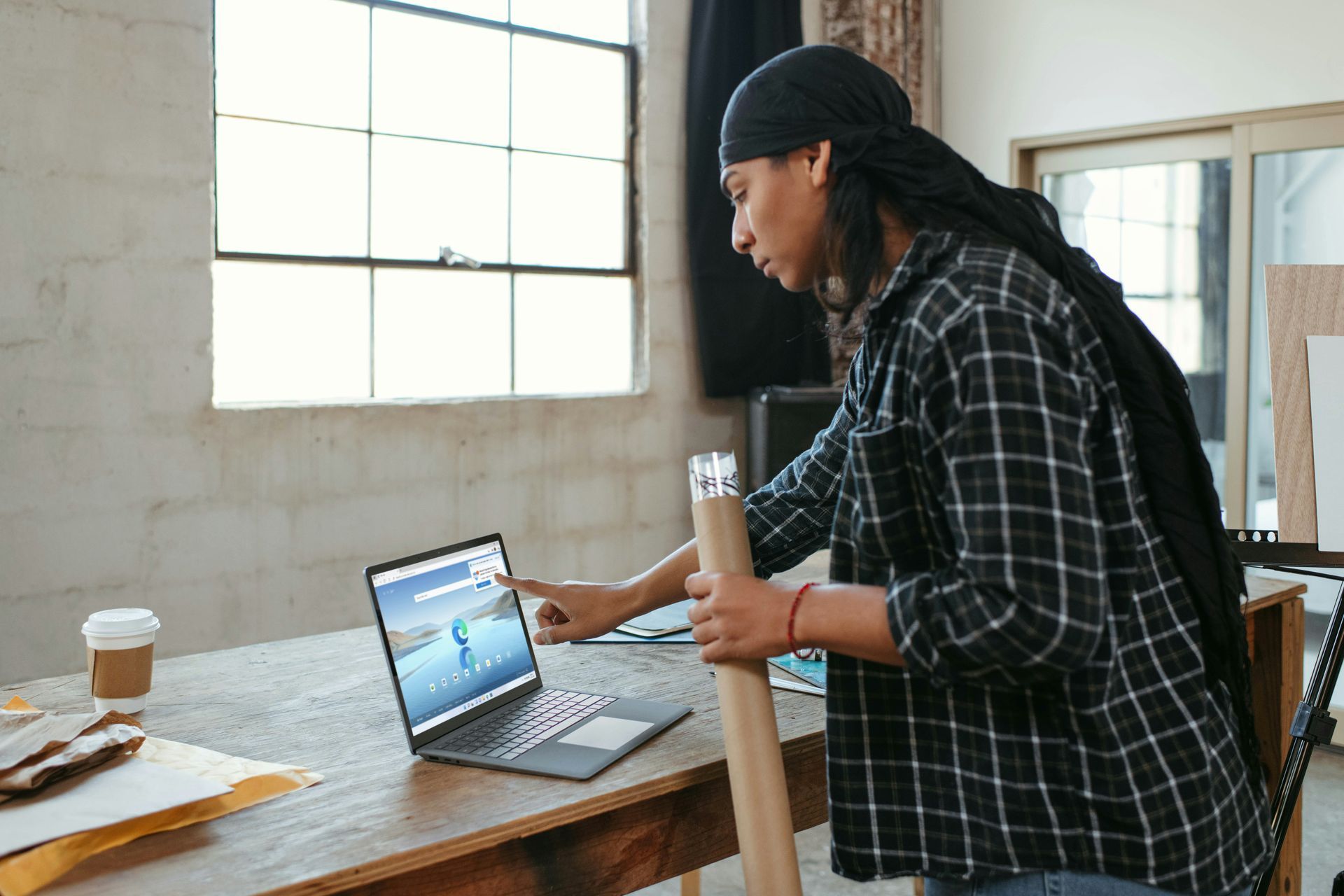 Person with dreadlocks in plaid shirt, using laptop, holding rolled paper in a bright room.