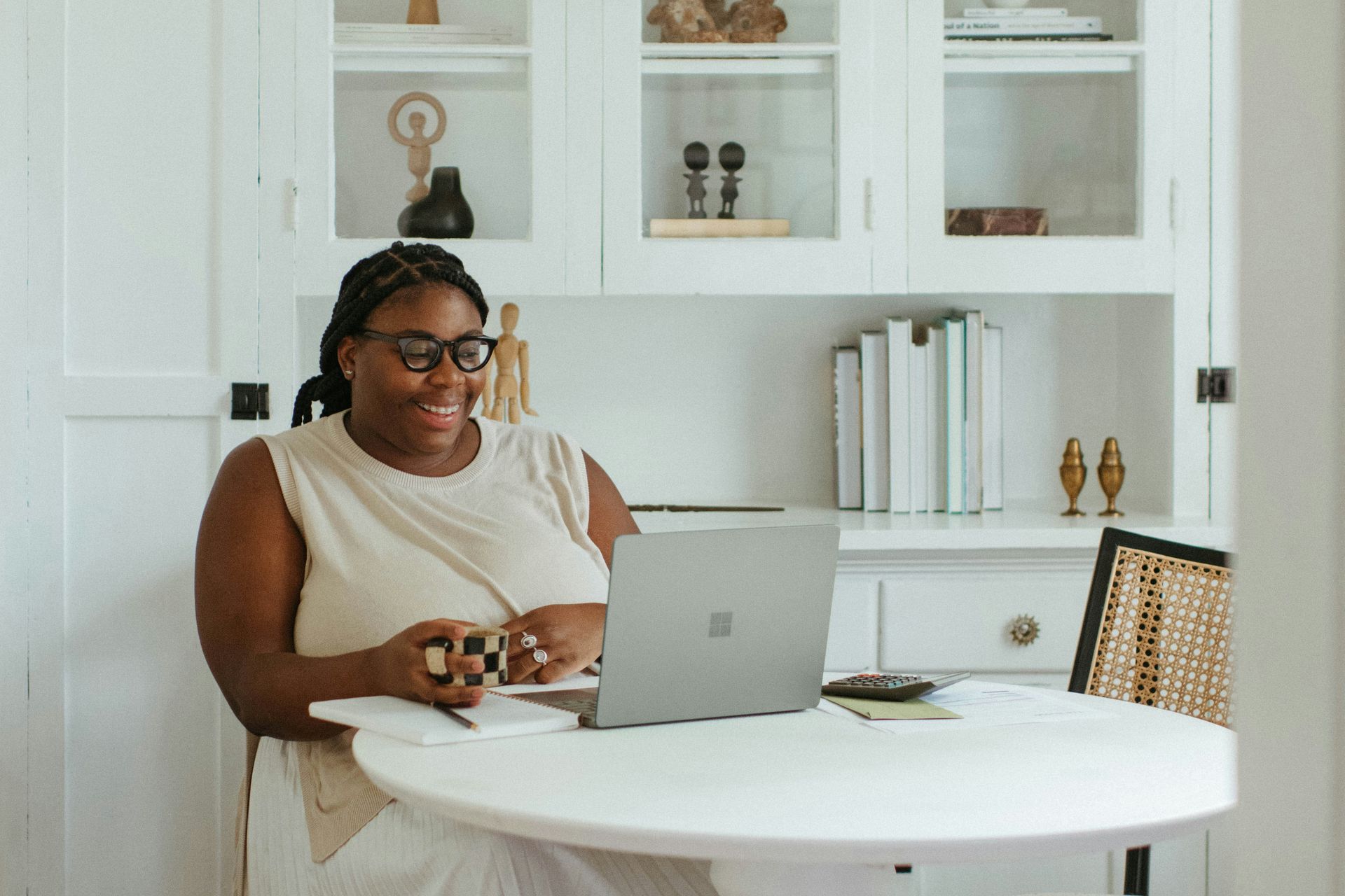 Woman in glasses smiles while on a laptop at a white table. A white cabinet is behind her.