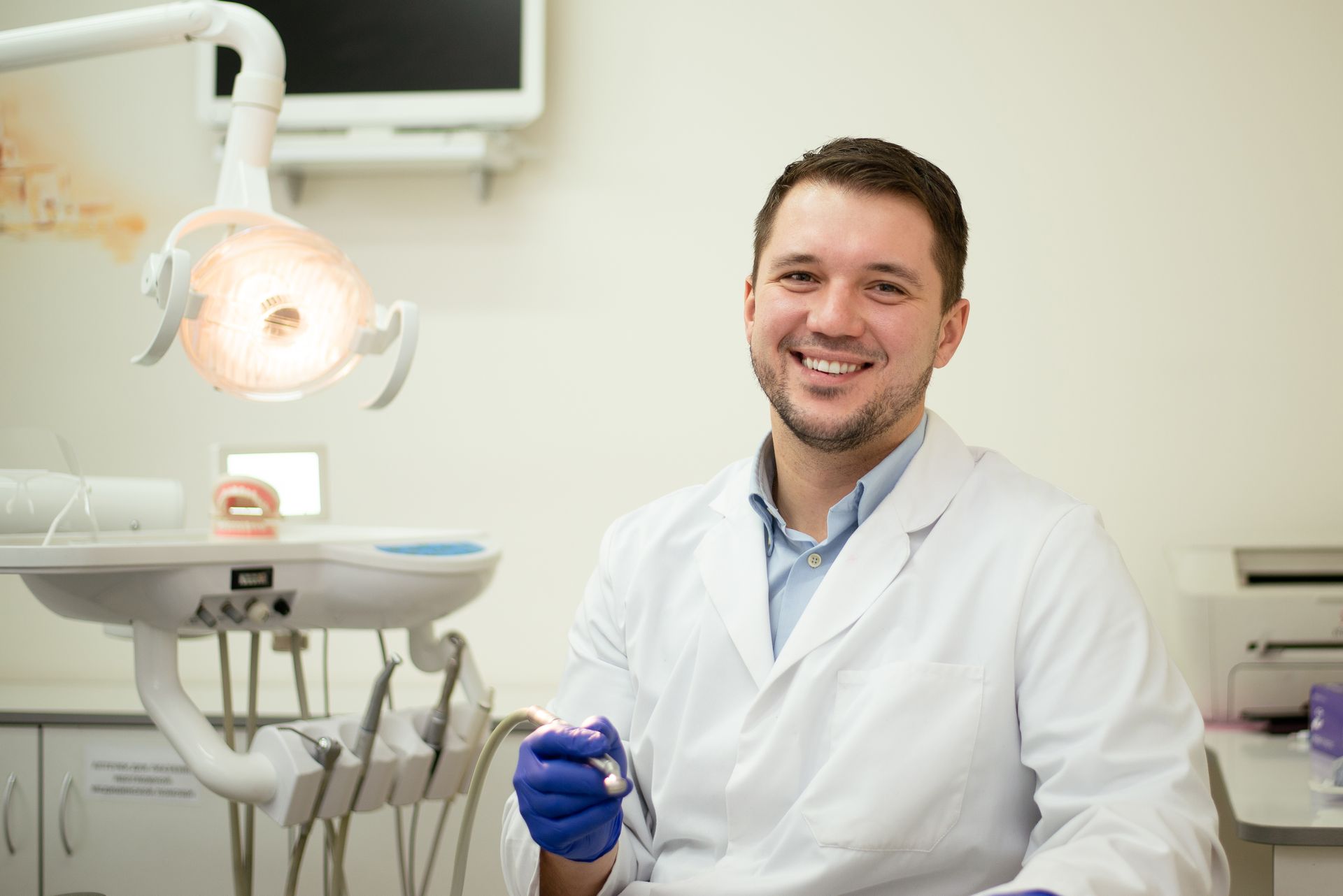 Dentist in white coat smiles at camera, dental tools and equipment visible in the office setting.