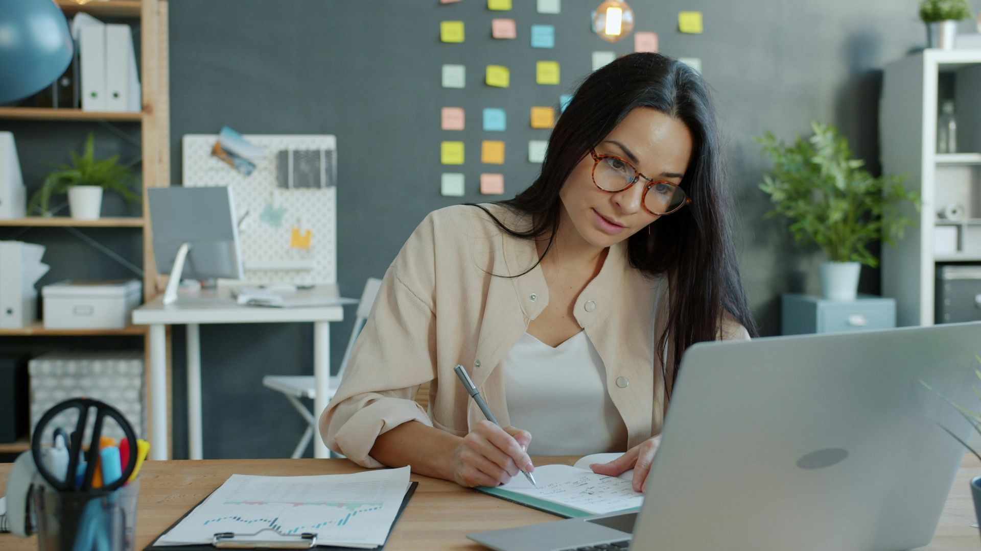 Woman with glasses writes in notebook at a desk with laptop, office setting, colorful sticky notes on wall.