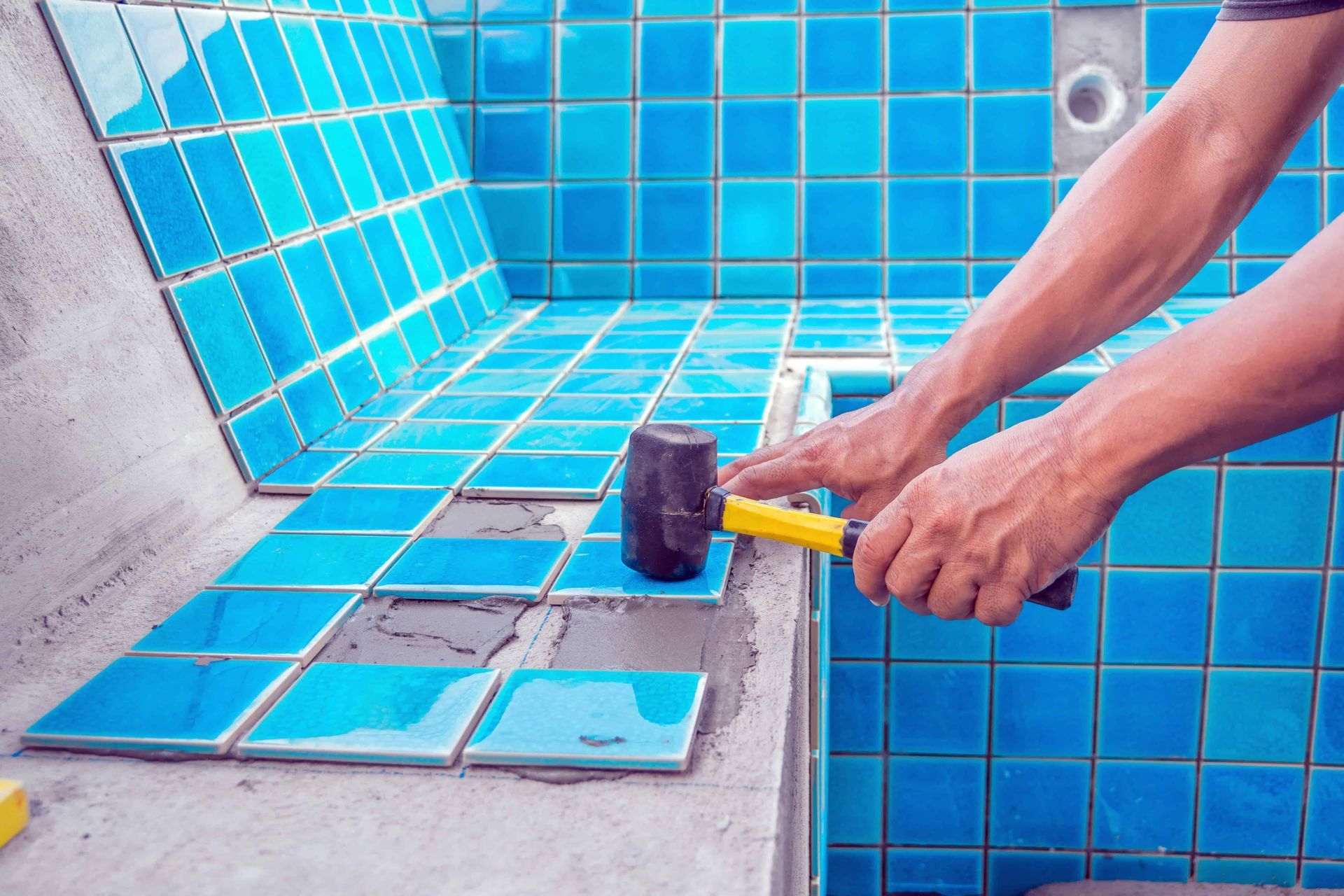 Person tiling a blue-tiled pool area, using a rubber mallet to position tiles.